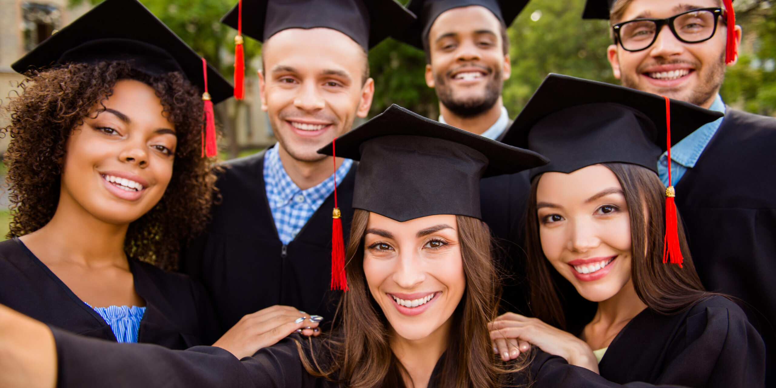 Selfie for memories. Six with cheerful graduates are posing for selfie shot, attractive brunette lady is taking, wearing gowns and mortar boards, outside on a summer day